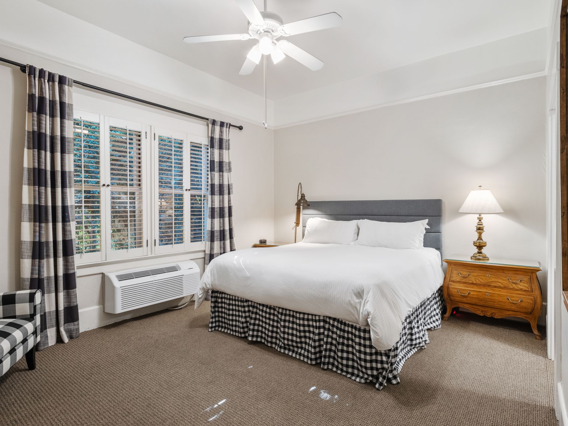 A bedroom with a king bed, grey headboard, buffalo plaid bed skirt, white bedding, windows with shutters, and wood dresser.