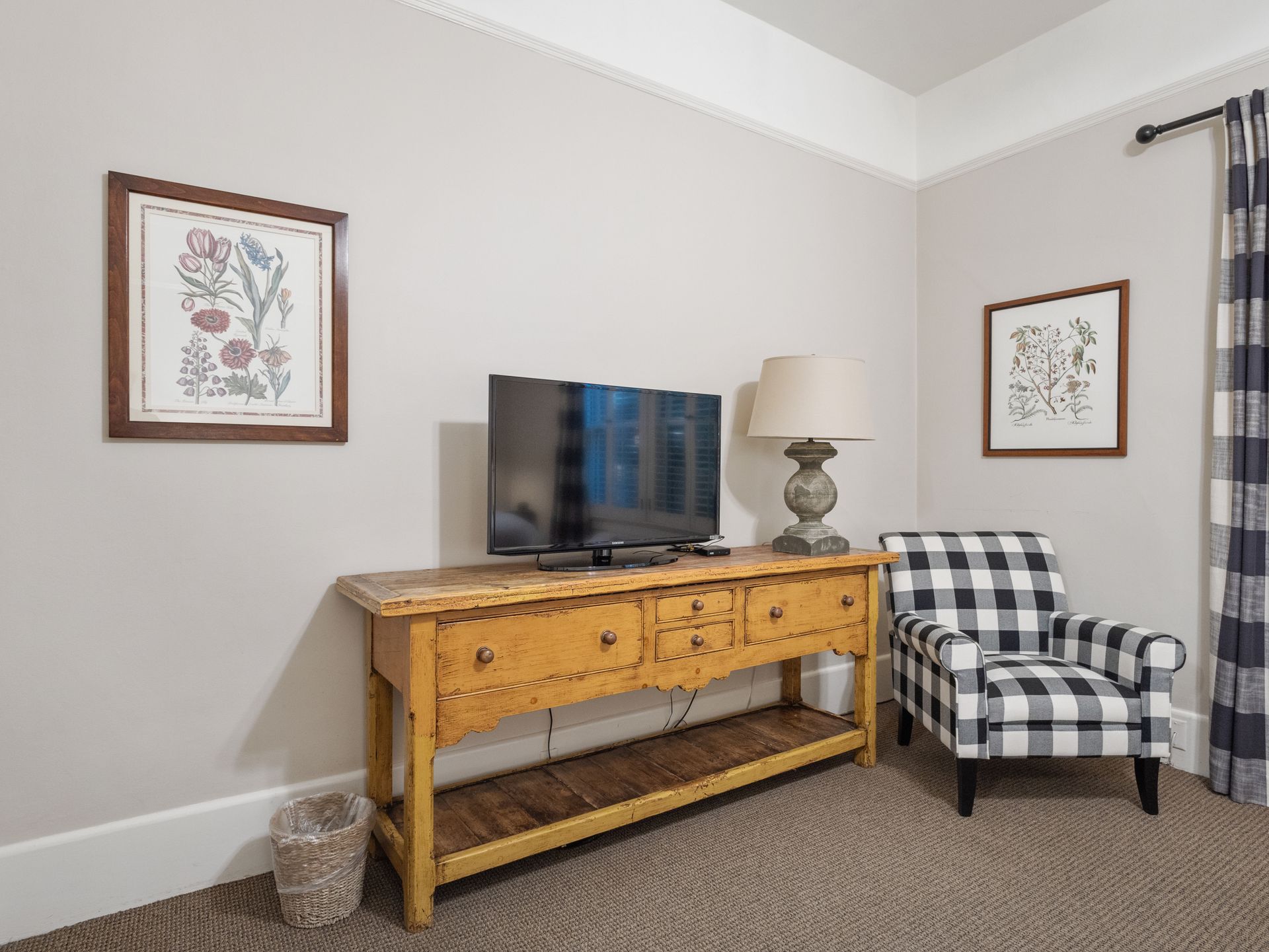 A TV on a long wooden console table next to a plaid armchair in a room with light walls and framed botanical prints.