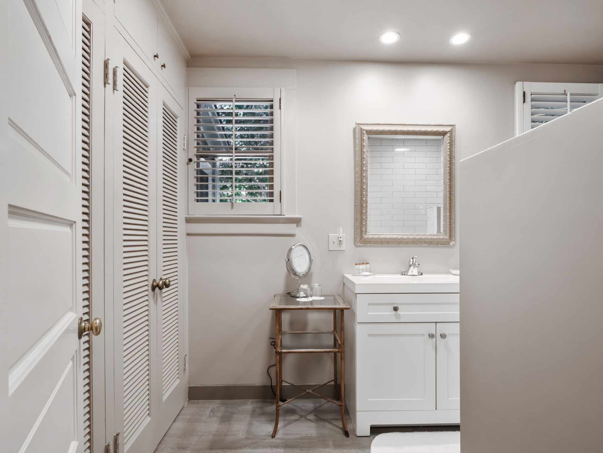 A bright bathroom with white louvered closet doors, a small metal vanity table, and a white sink under a framed mirror.