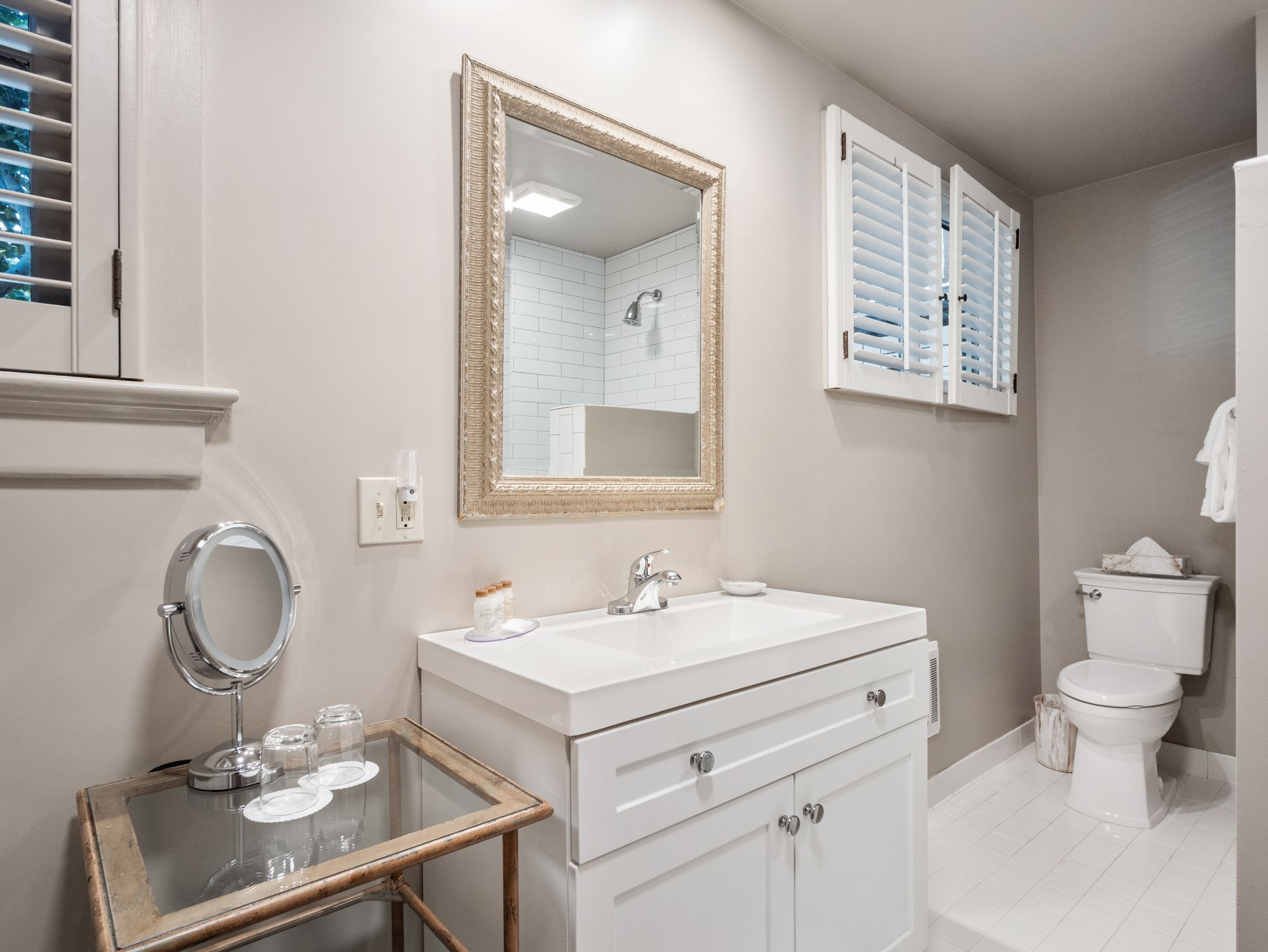 A bathroom featuring a white vanity with a mirror, a small glass side table with a vanity mirror, and a toilet.