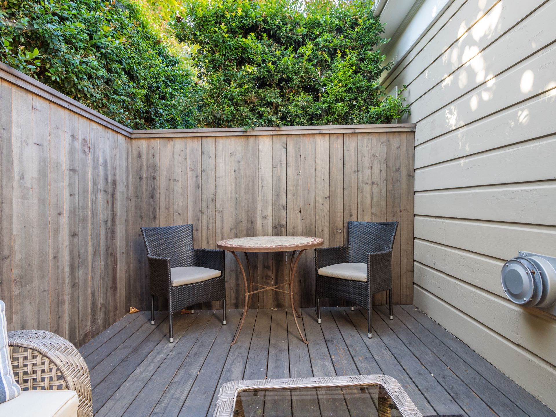 Two wicker chairs and a small round table on a wooden deck next to a cedar fence, enclosed by lush greenery.
