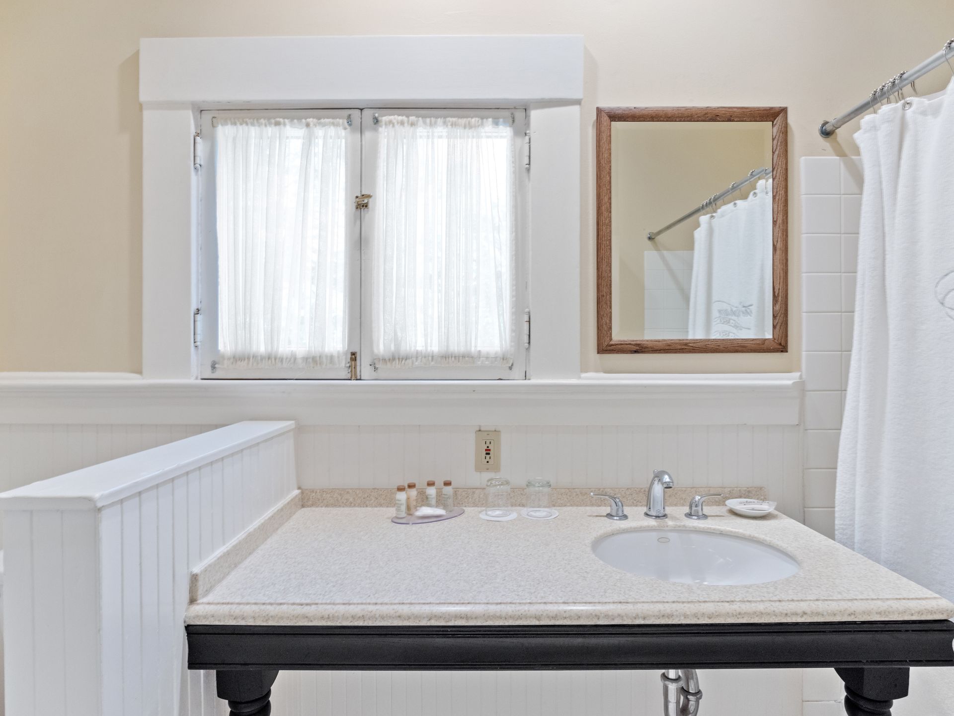 A bathroom vanity with a sink, toiletries, mirror, and a window with white curtains next to a shower curtain.