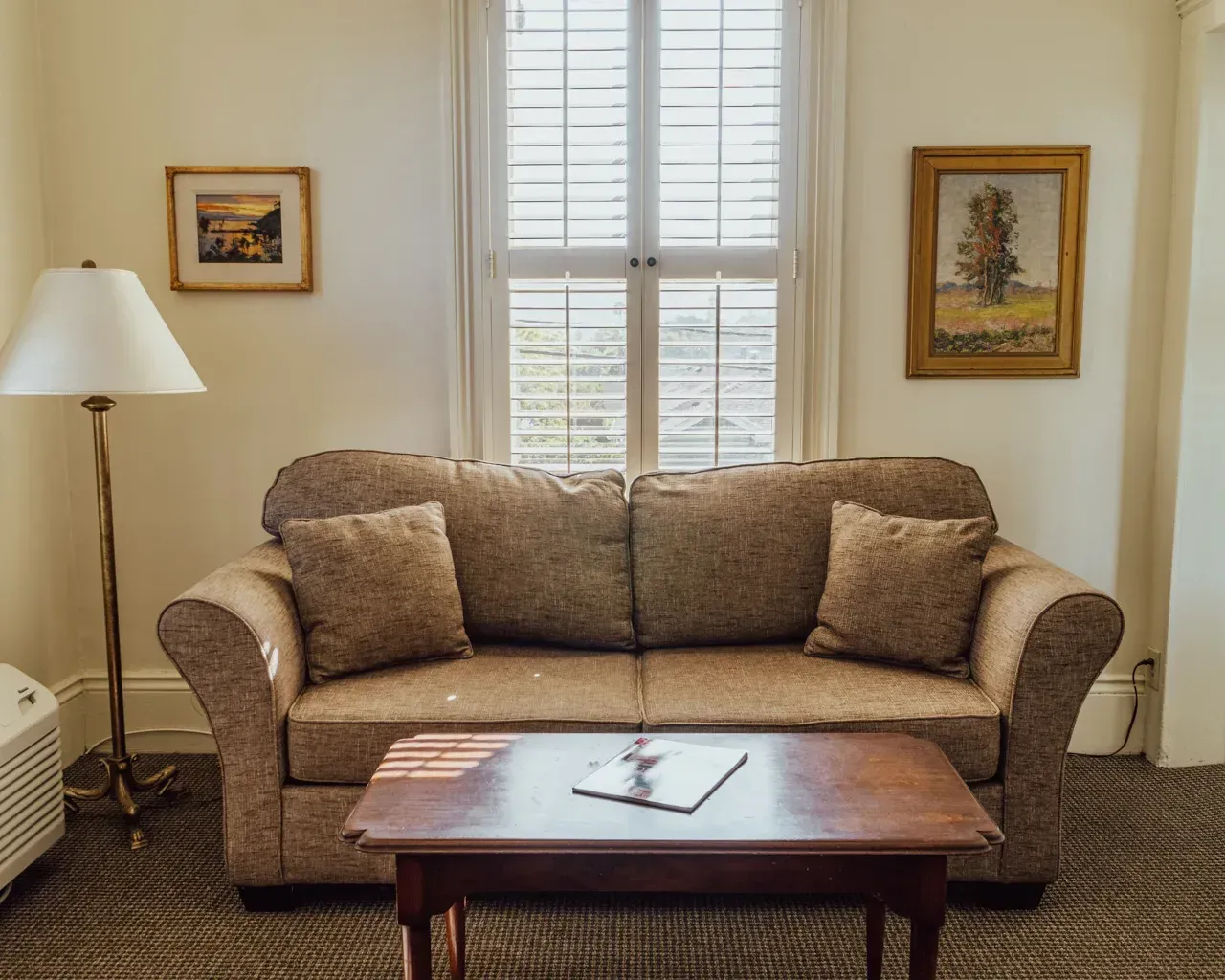 A tan sofa with two cushions and accent pillows sits before a window with shutters, with a coffee table and floor lamp.