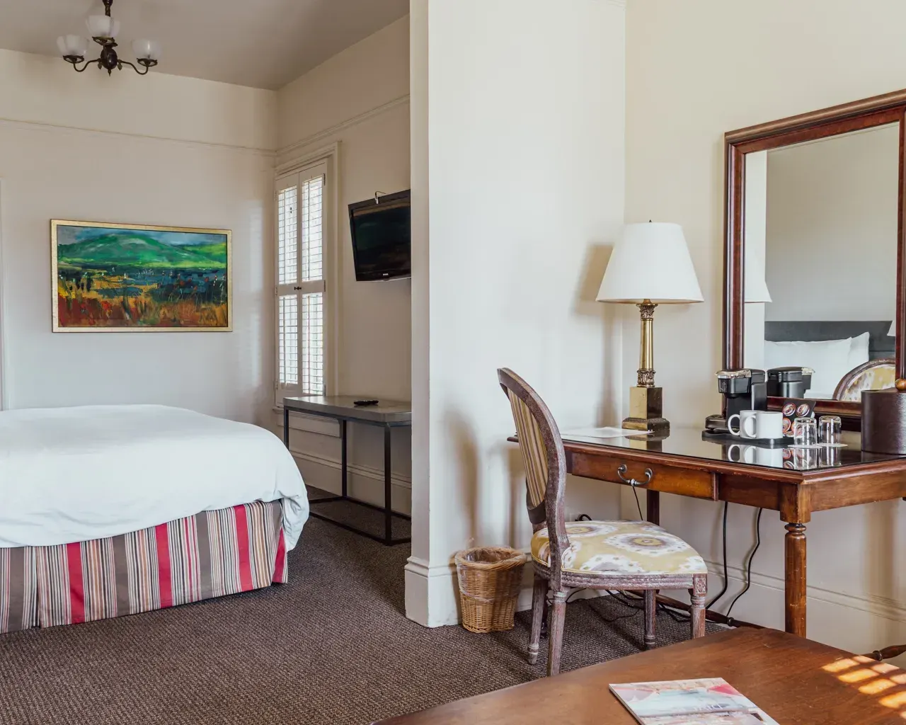 A bedroom with a bed, a patterned bedskirt, a landscape painting, a desk with a lamp, a chair, and a vanity mirror.