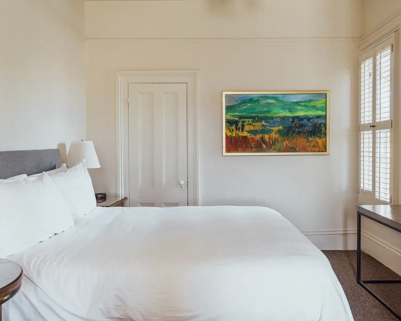 A white-walled bedroom featuring a bed with white linens, a framed landscape painting, a closed door, and window shutters.