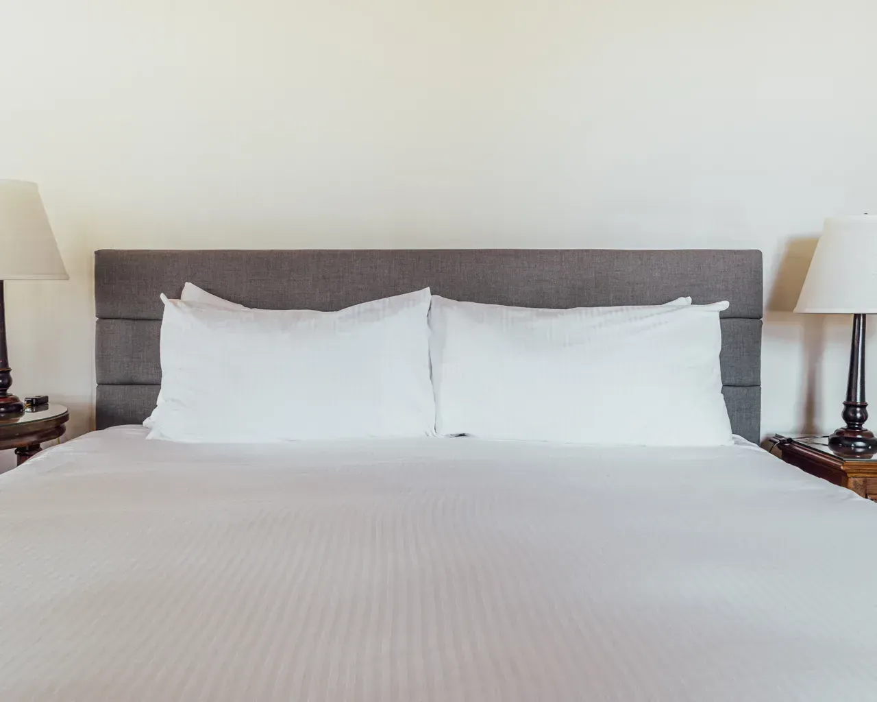A neatly made bed with white bedding and two white pillows against a gray fabric headboard, flanked by two lamps.