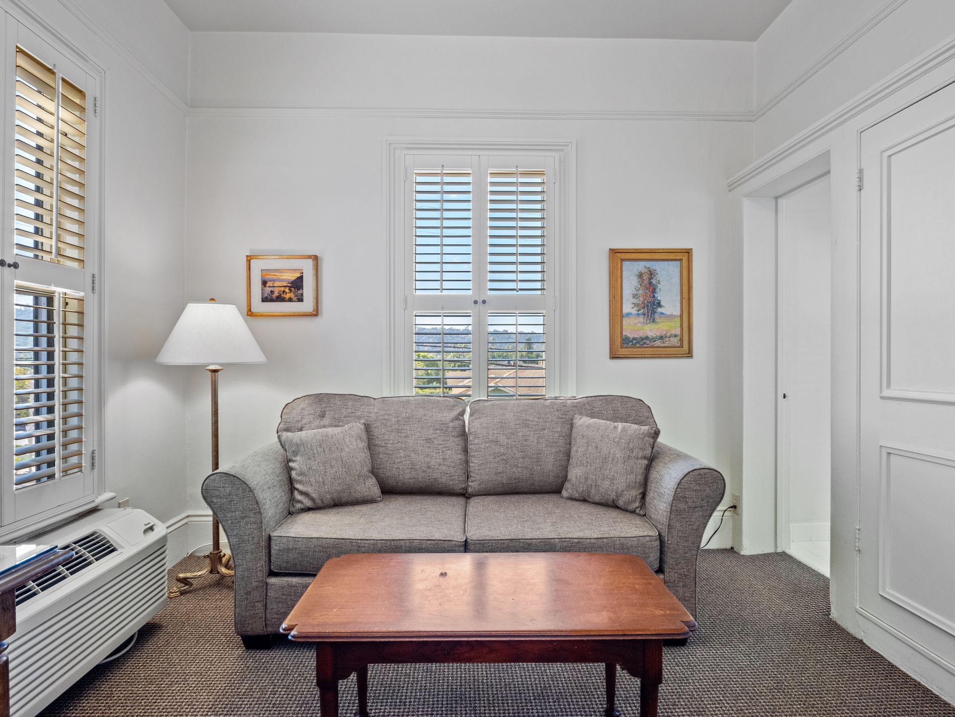 A gray sofa sits in front of a shuttered window, flanked by a floor lamp and two framed paintings in a white room.