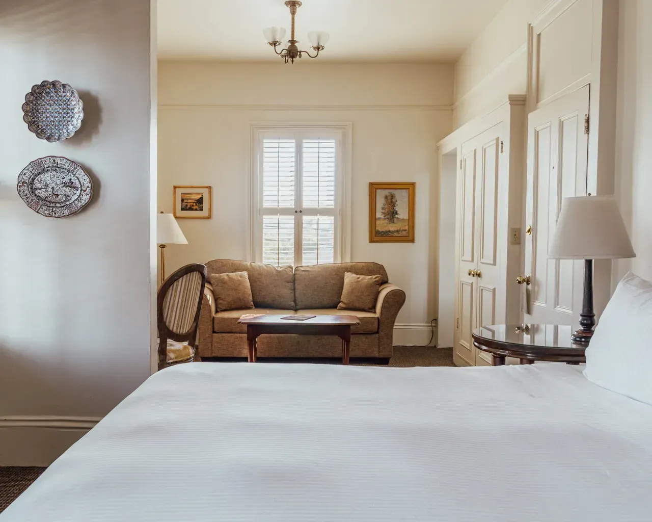 A hotel room featuring a white bed in the foreground, facing a sitting area with a sofa, a coffee table, and a window.