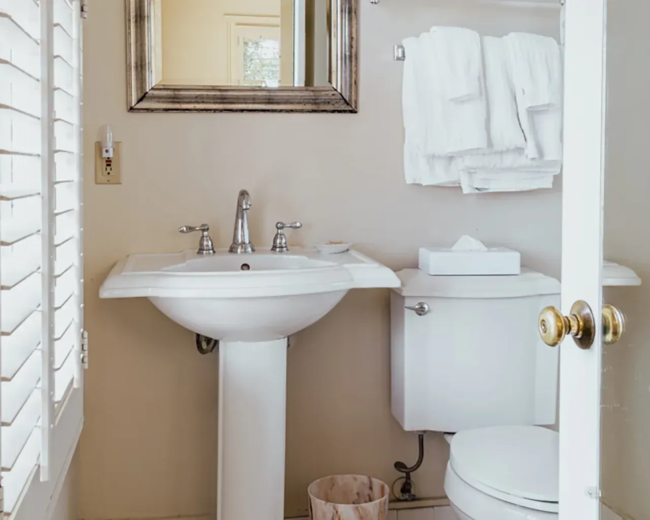 A pedestal sink with silver fixtures and a mirror above it, next to a white toilet with towels in a neutral-toned bathroom.