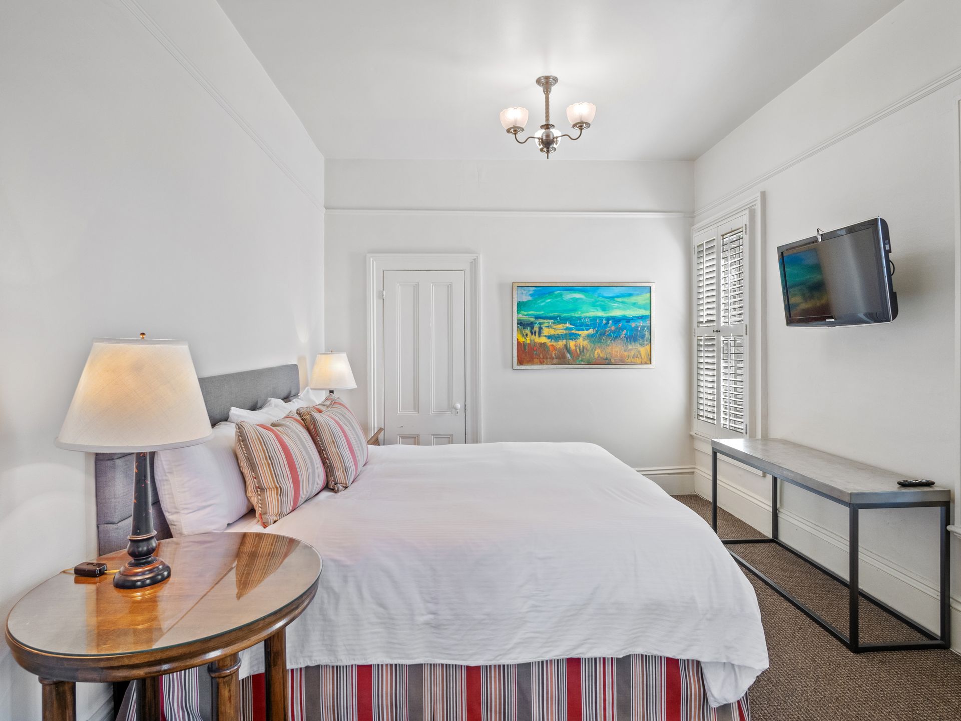A bedroom with a bed, a round wooden nightstand with a lamp, a wall-mounted TV, and a landscape painting.