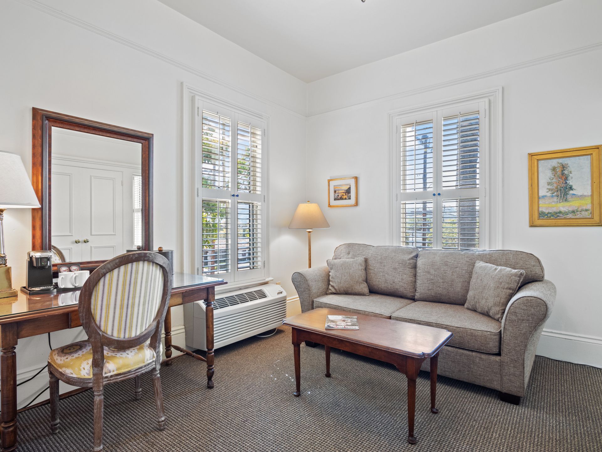 A hotel room featuring a sofa, coffee table, desk with a chair and mirror, two windows with shutters, and wall art.