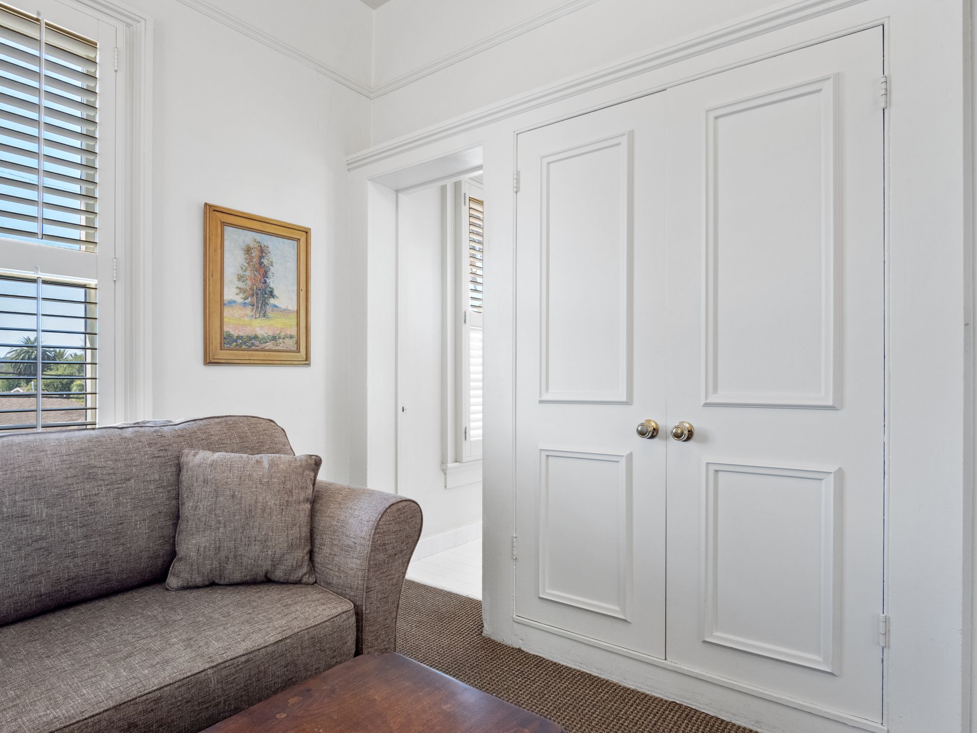 A grey sofa sits near a white double door and a window with blinds in a room featuring a landscape print on the wall.