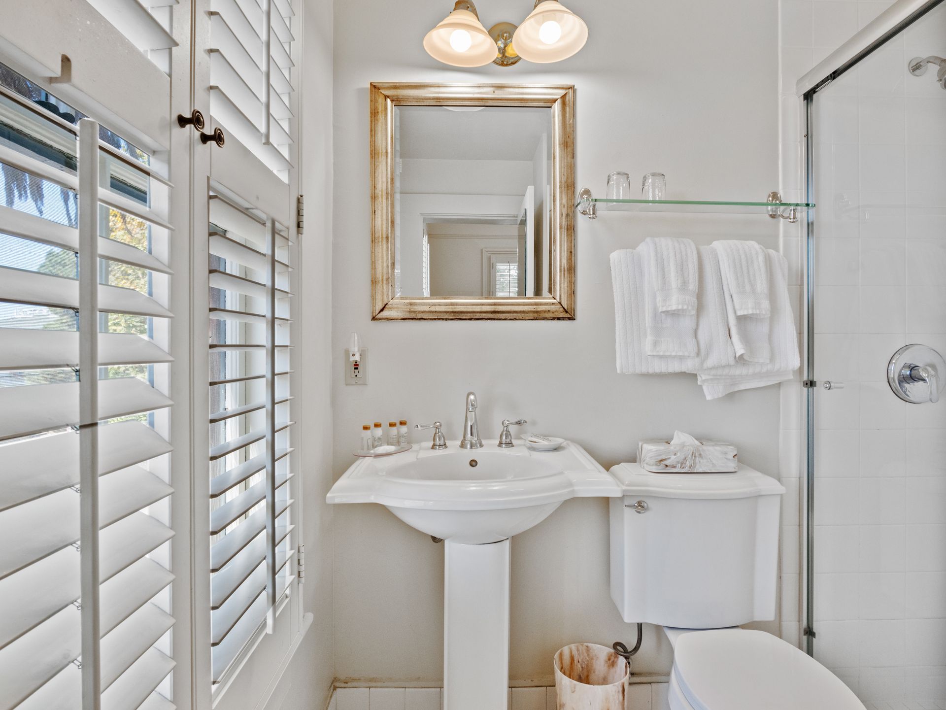 A well-lit, white-walled bathroom with a pedestal sink, framed mirror, wall sconces, towels on a rack, and a toilet.
