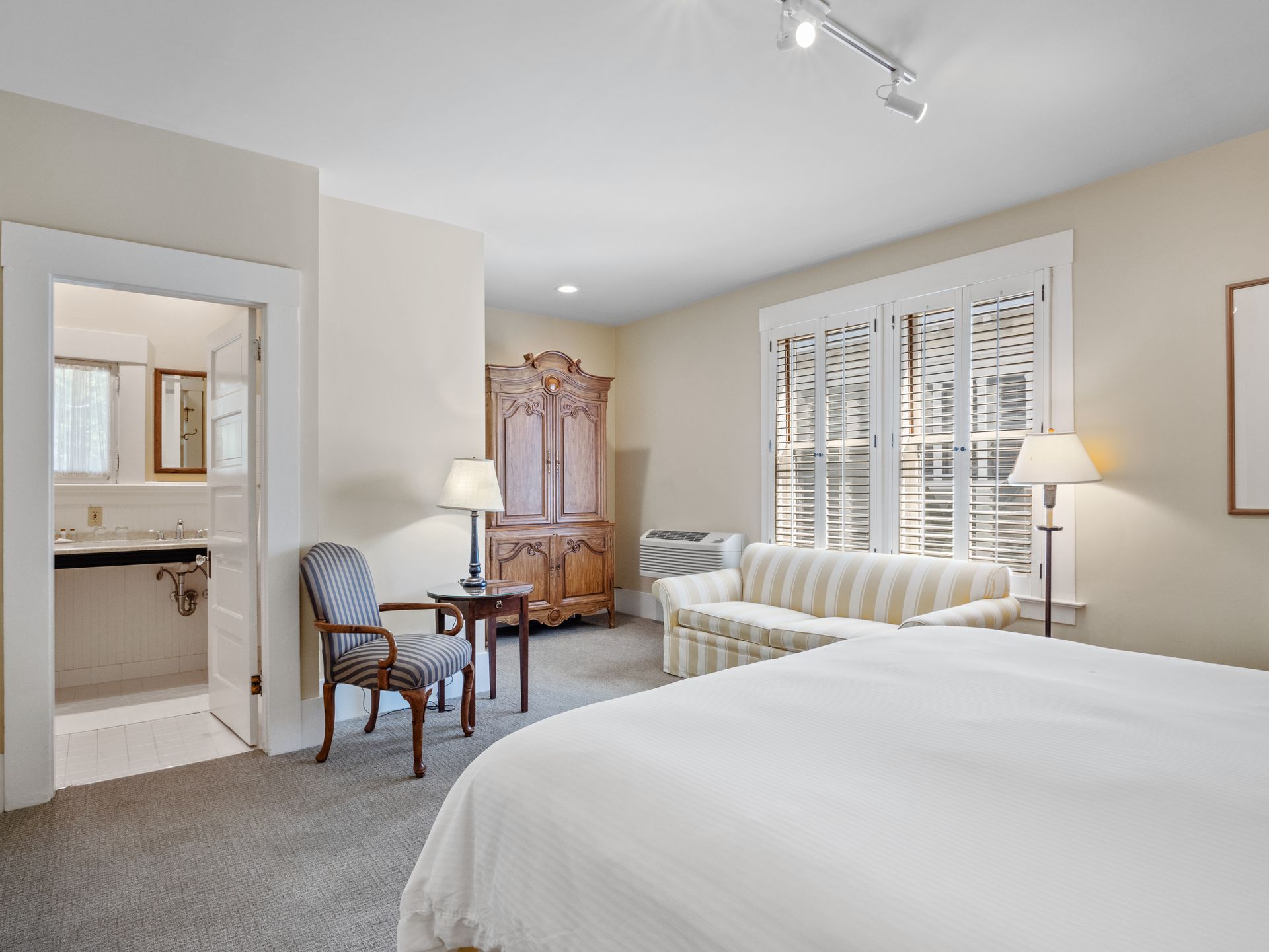 A hotel room with a large bed in the foreground, a striped sofa, an armchair, an armoire, and an open door to a bathroom.