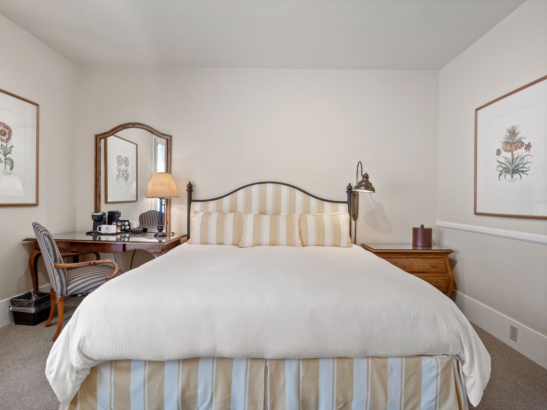 A hotel room featuring a queen bed with striped pillows, a wooden desk with a lamp, and framed botanical art on the walls.
