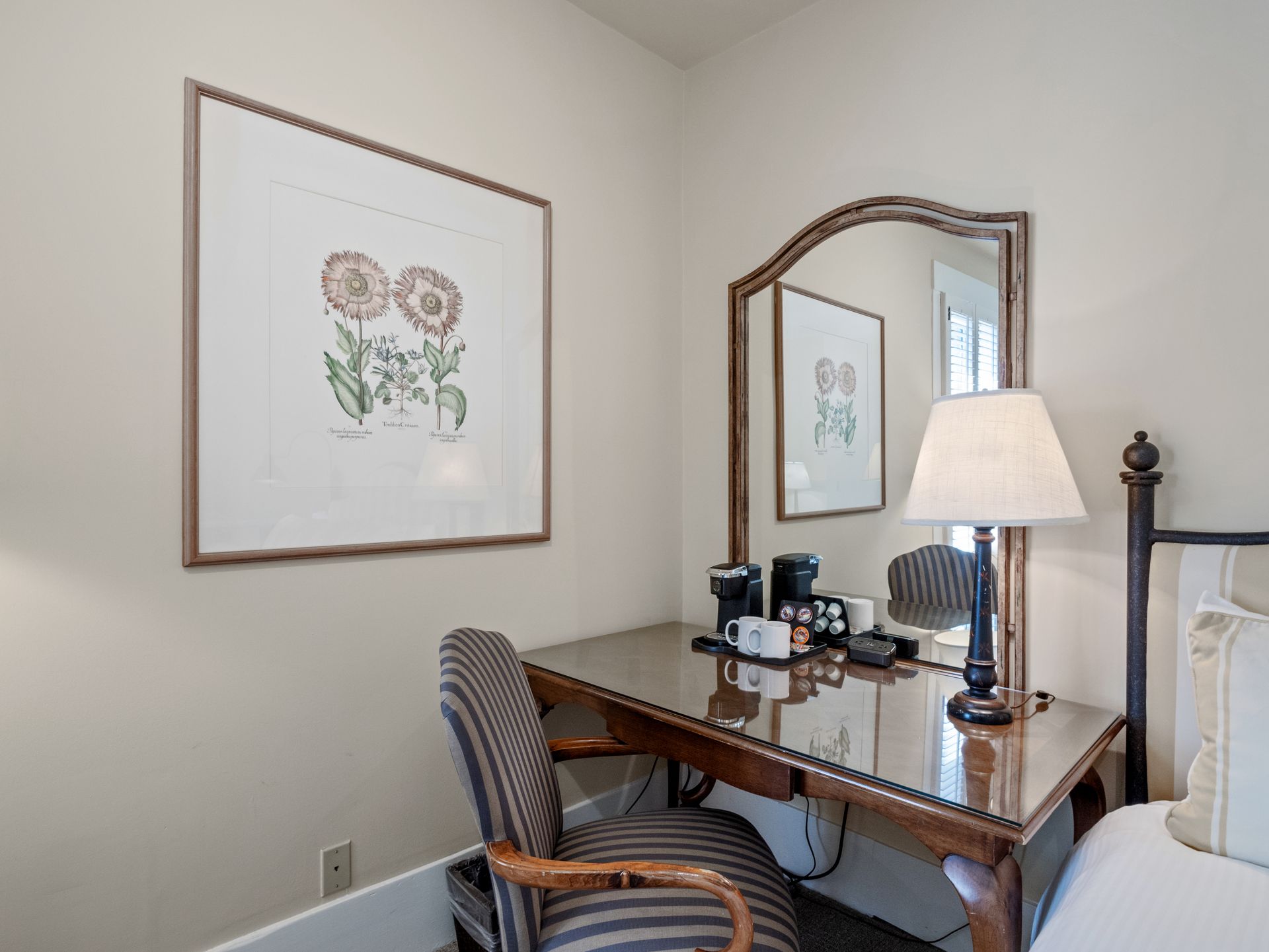 A wooden desk and striped chair sit against a wall featuring botanical artwork, near a mirror and a bedside lamp.