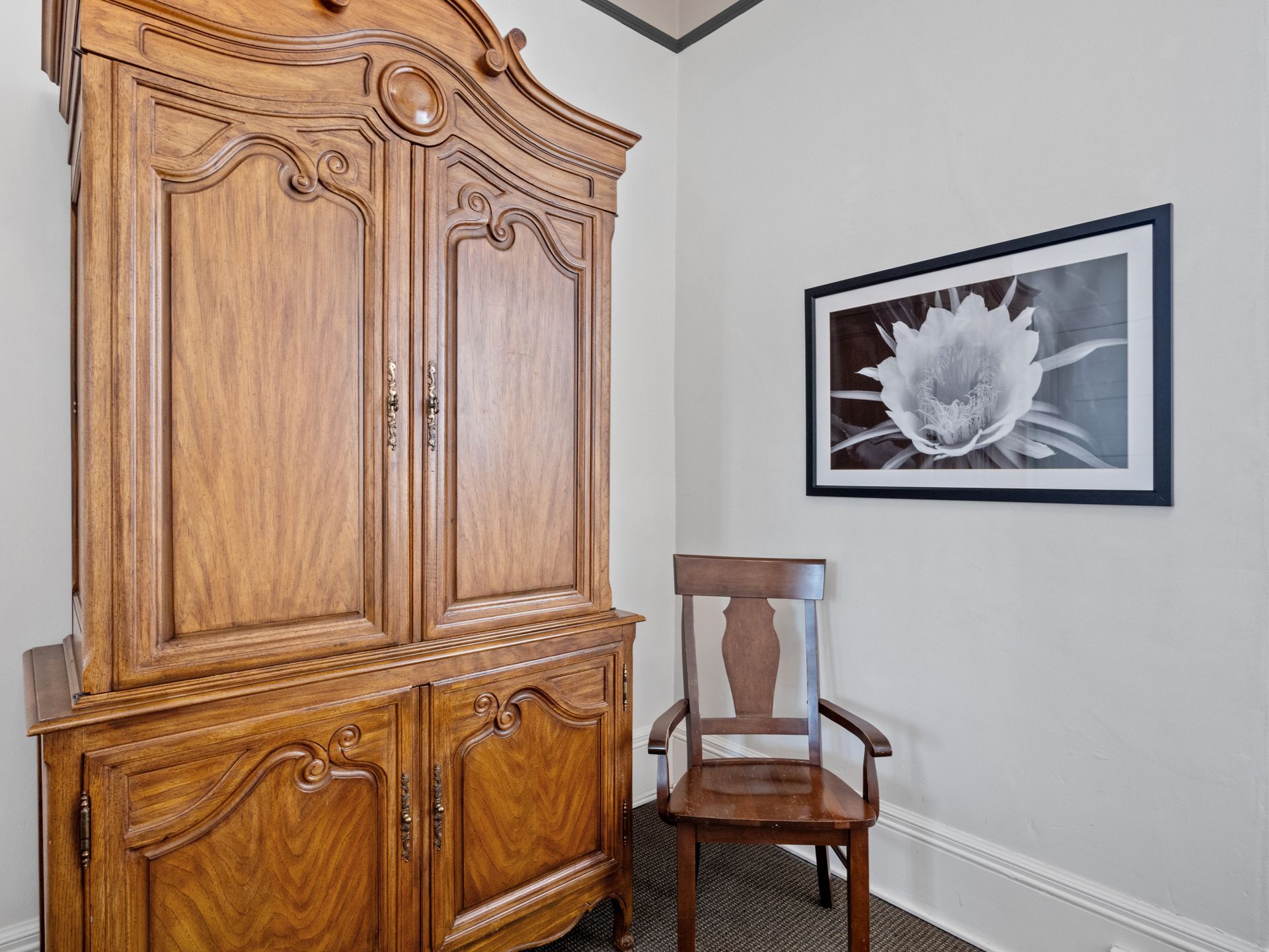 A large wooden armoire stands next to a wooden chair in a room with a framed black-and-white floral print on the wall.