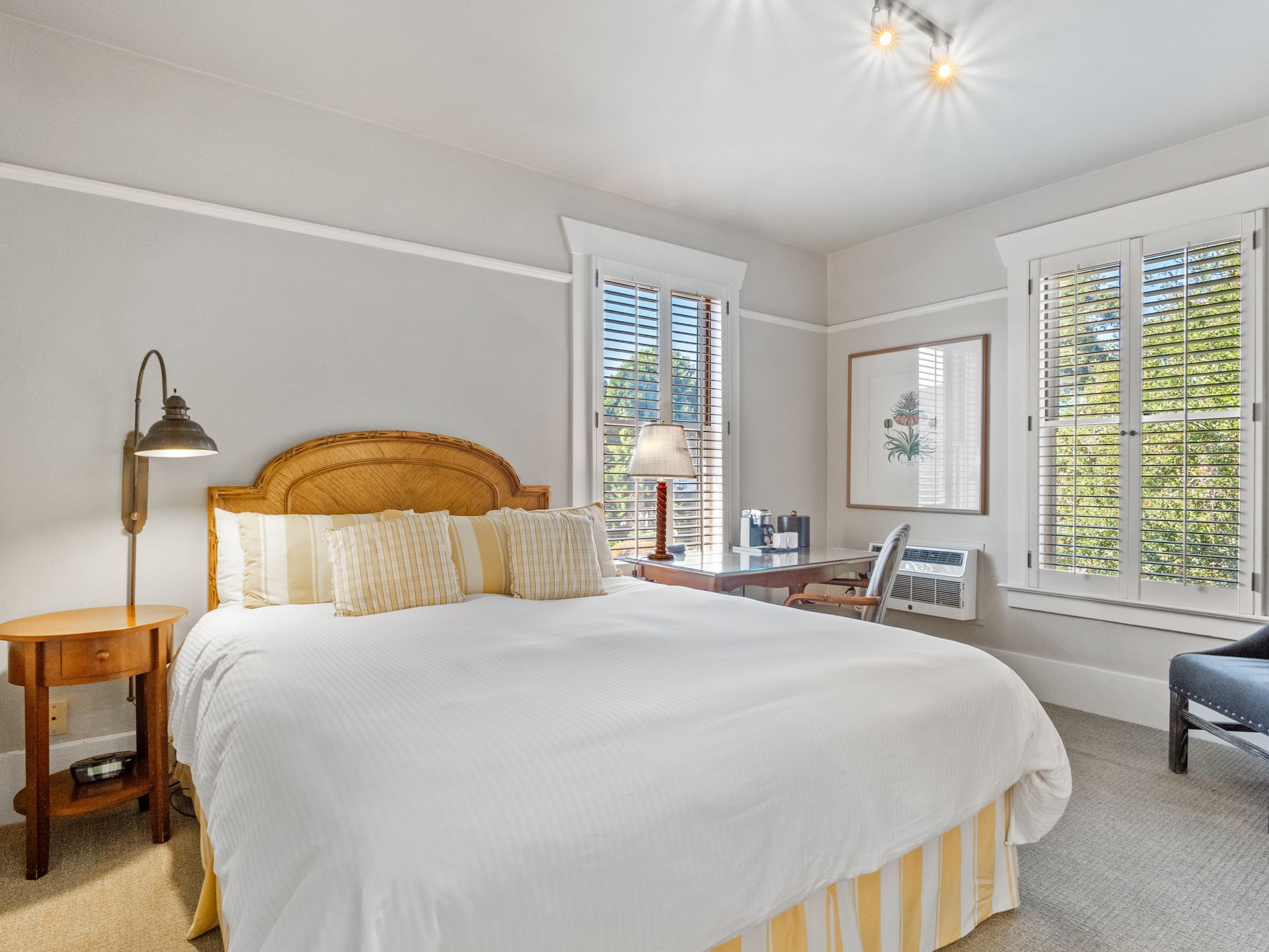 A bright bedroom with a king-sized bed, white linens, a wooden nightstand with a lamp, and windows with white shutters.