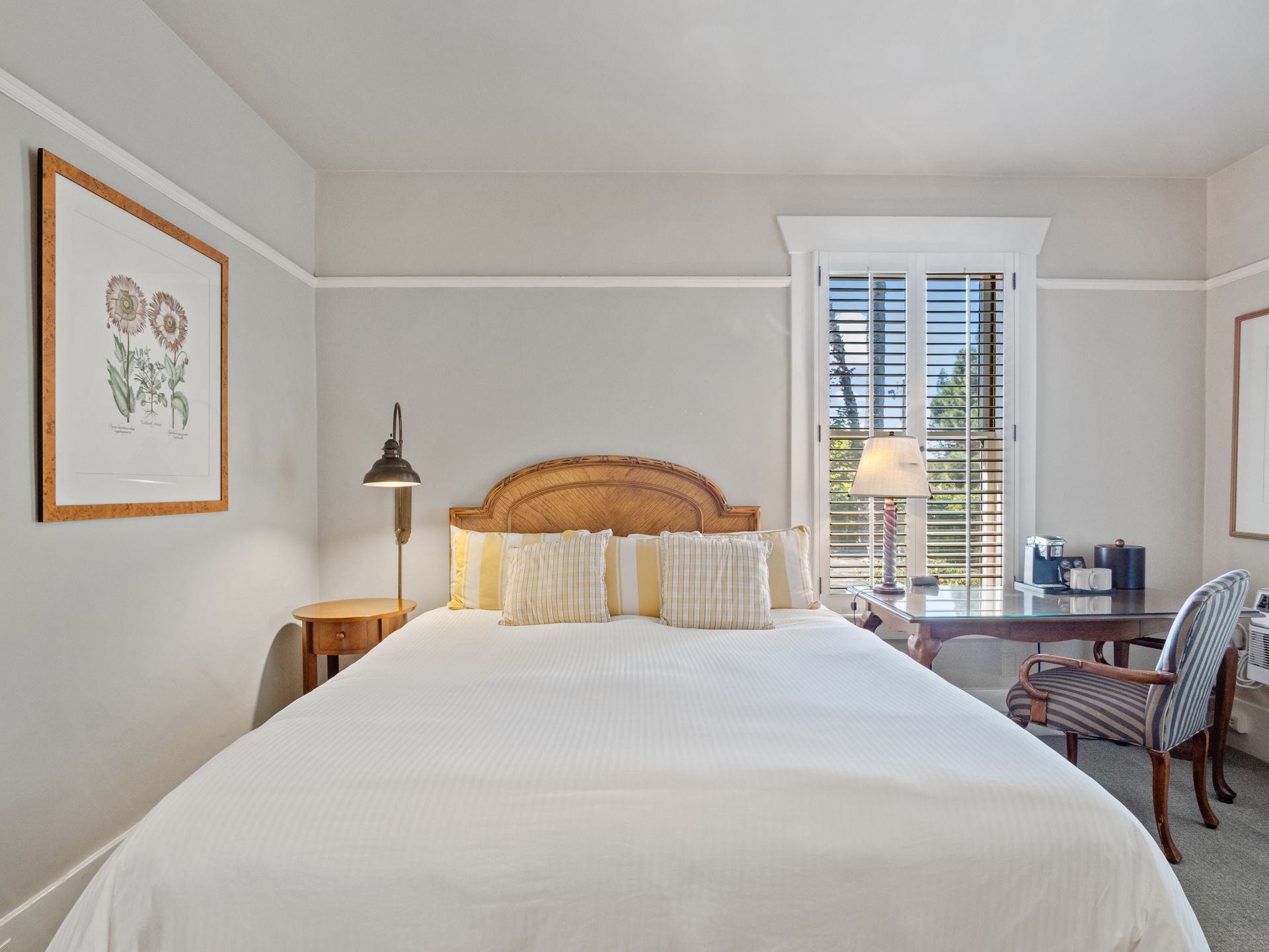 A bedroom with a queen bed, beige headboard, white bedding, a desk with a chair, and framed floral artwork on the wall.