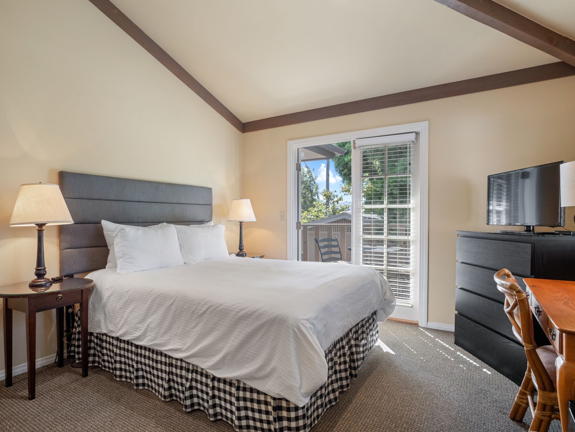 A hotel room with a queen bed, gray headboard, two nightstands, a television on a dresser, and a sliding glass door.