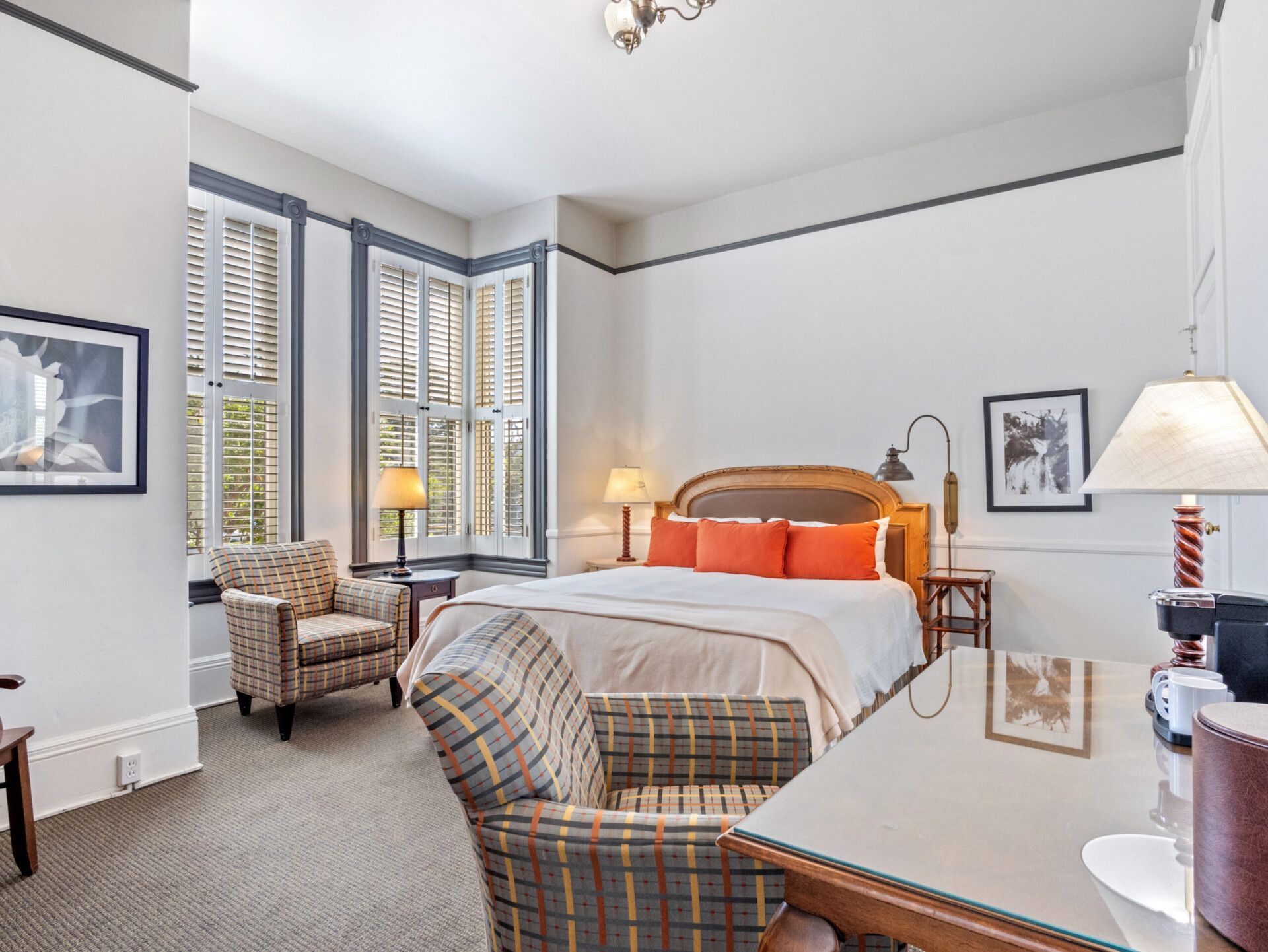 A hotel bedroom with a bed featuring orange pillows, two patterned armchairs, a glass-topped desk, and shuttered windows.