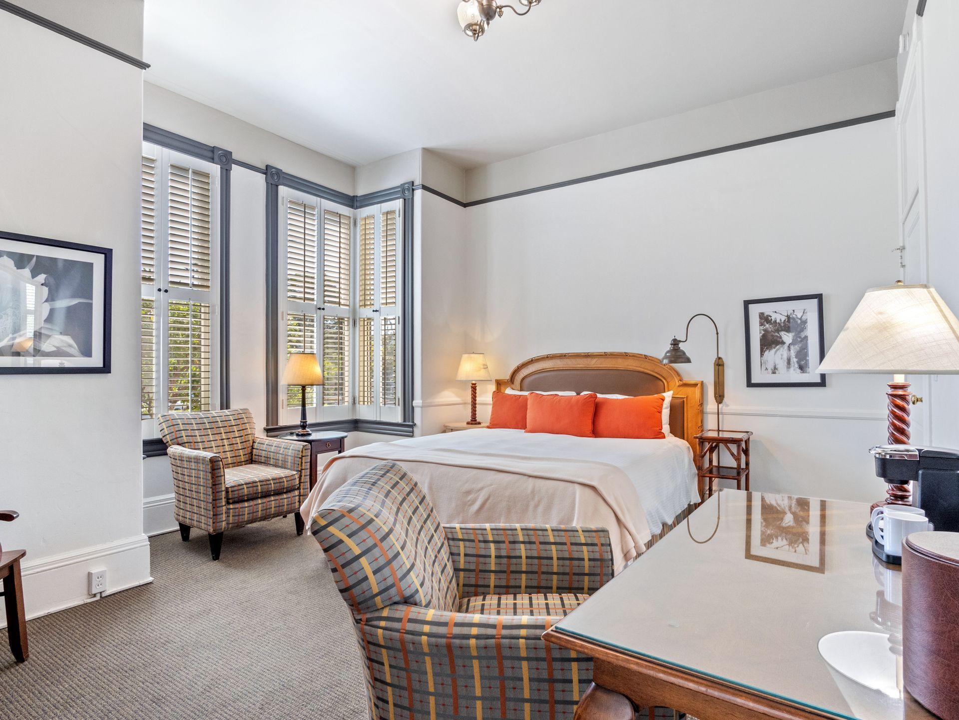 A bedroom featuring a bed with orange pillows, two plaid armchairs, a glass-topped desk, and white walls with trim.