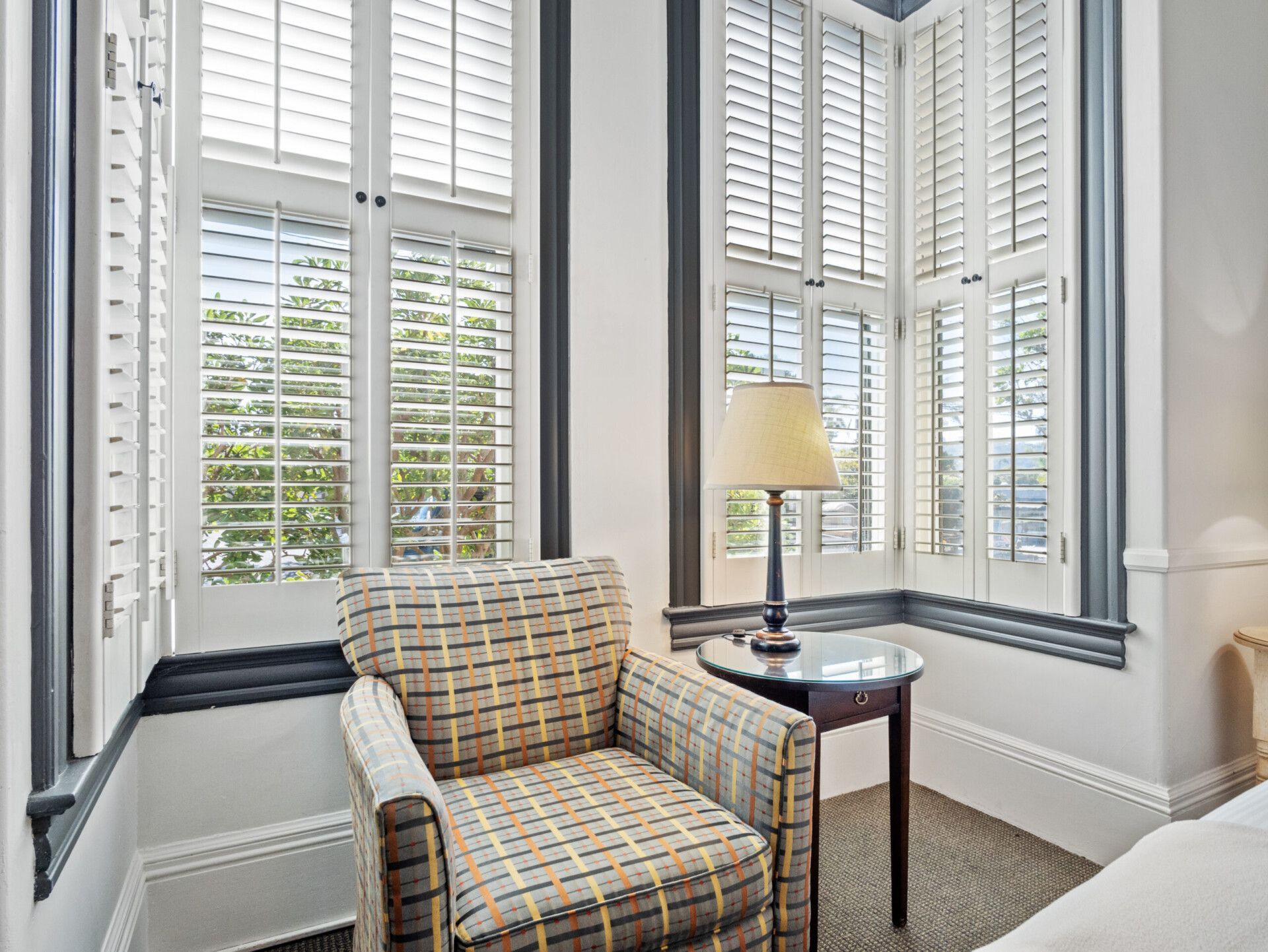 A patterned armchair and a side table with a lamp sit in a corner beside tall windows with white plantation shutters.