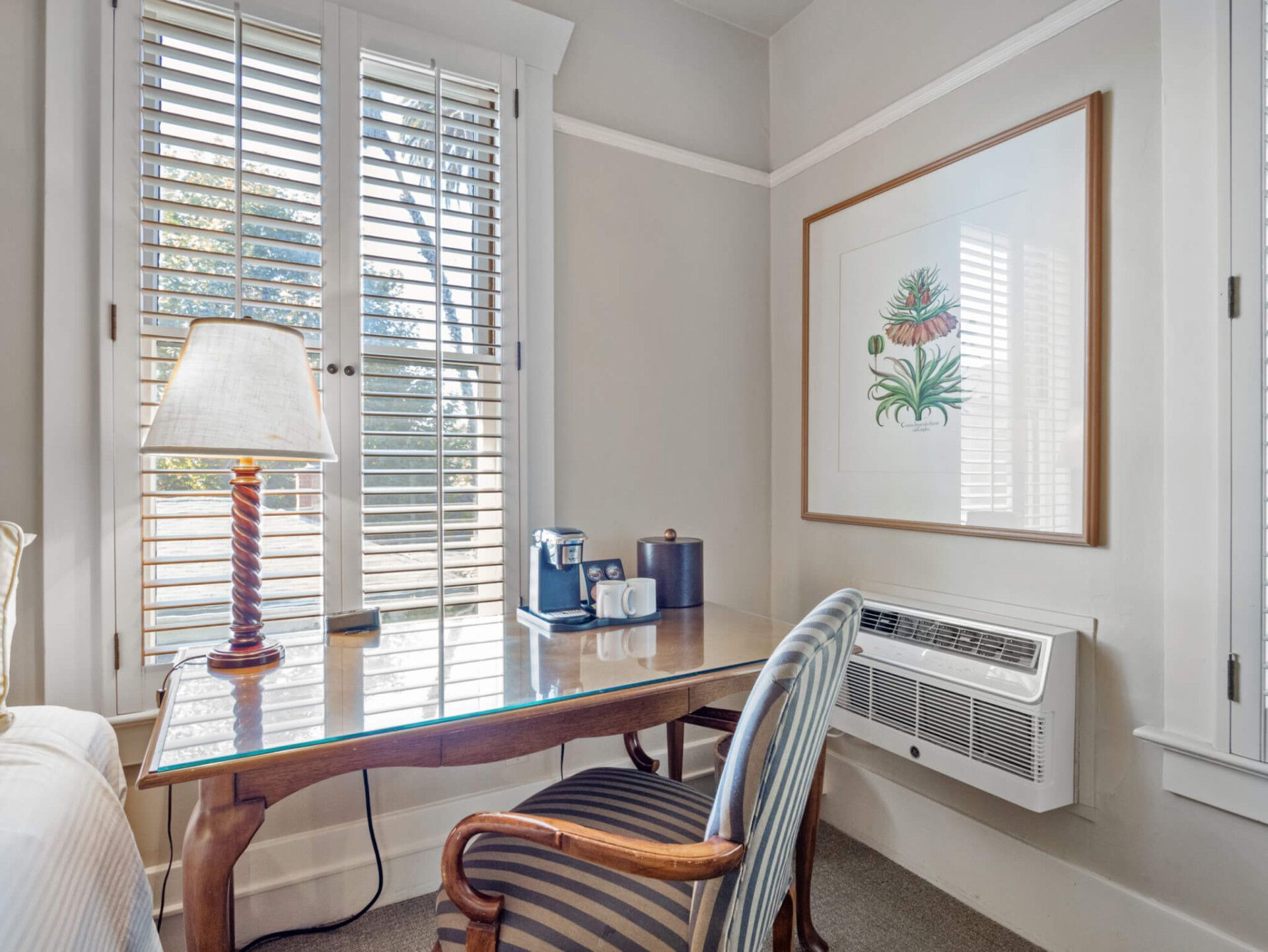 A desk with a lamp, coffee maker, and chair beside a window with plantation shutters and a wall-mounted air conditioner.
