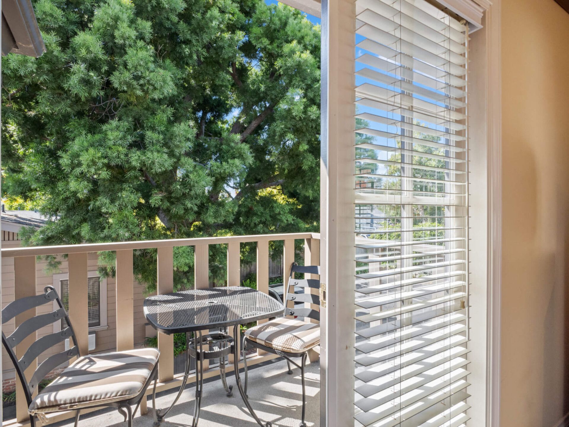A patio balcony with a small table and two chairs, viewed from inside through a glass sliding door with white blinds.