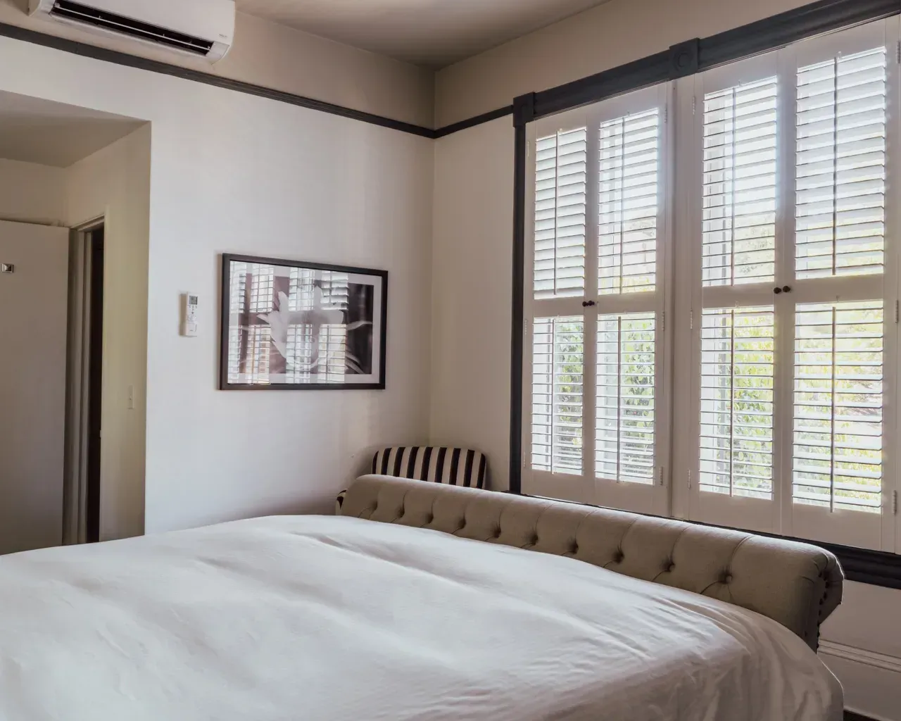 A tidy bedroom featuring a tufted beige bed, plantation shutters, and a framed black-and-white print on a neutral wall.
