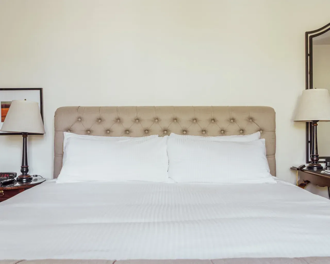 A neutrally colored, tufted headboard on a bed with white pillows and bedding, flanked by two lamps on side tables.
