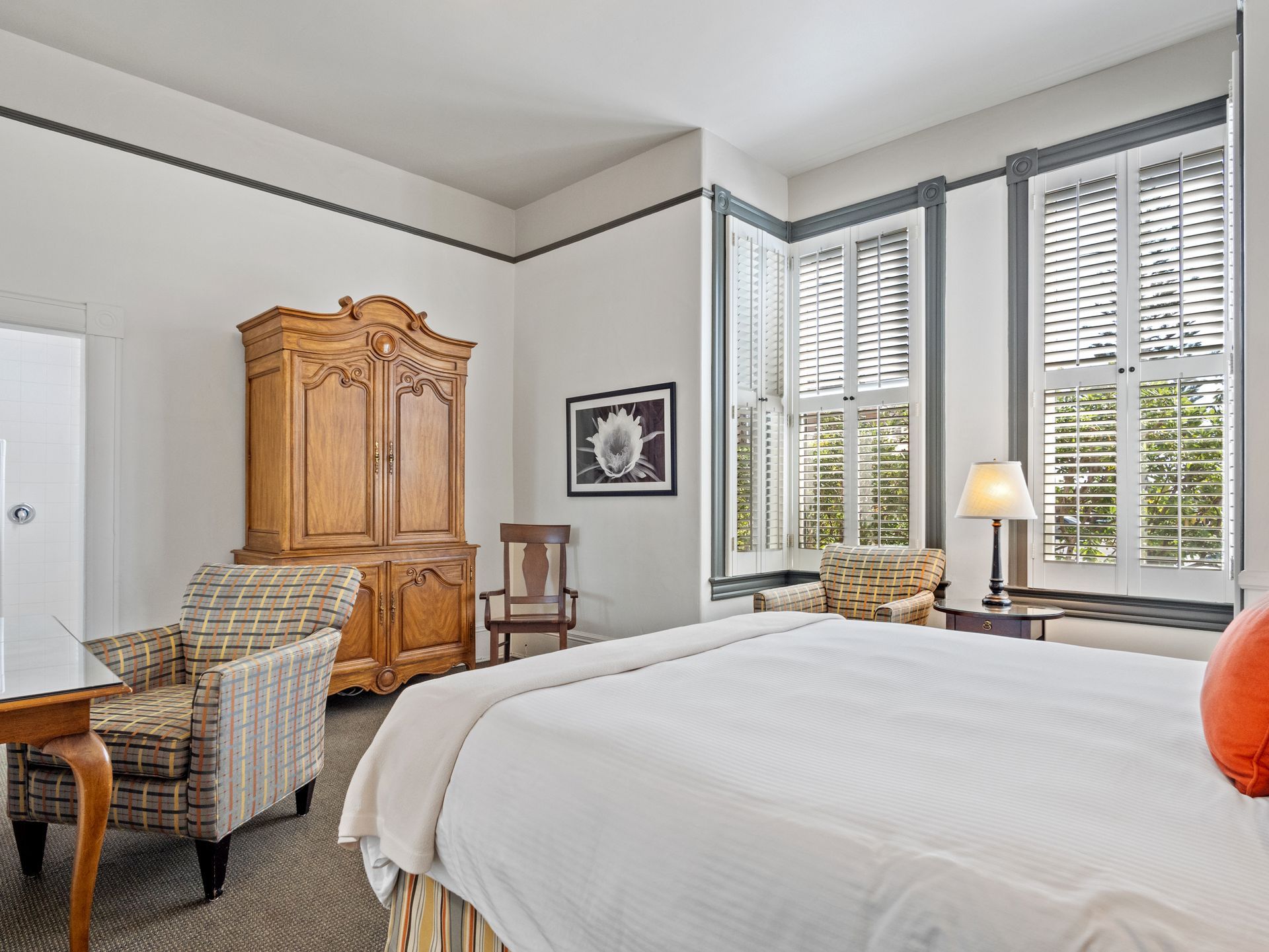 A hotel room featuring a bed with white bedding, a large wooden armoire, an armchair, and windows with white shutters.