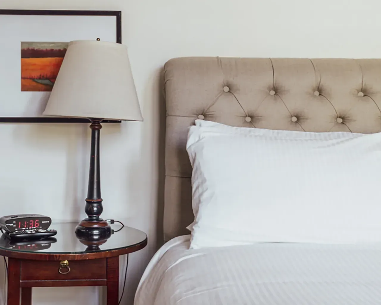 A beige tufted headboard, a white pillow, a small wooden nightstand with a lamp, and a digital clock next to a wall art.