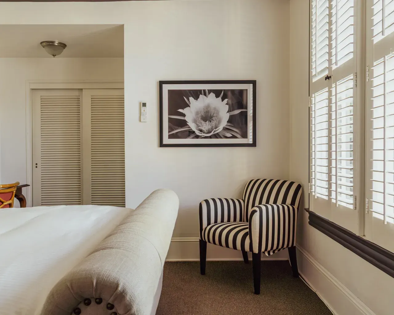 A black and white striped armchair in a corner next to a window with white shutters, near a bed and framed floral art.
