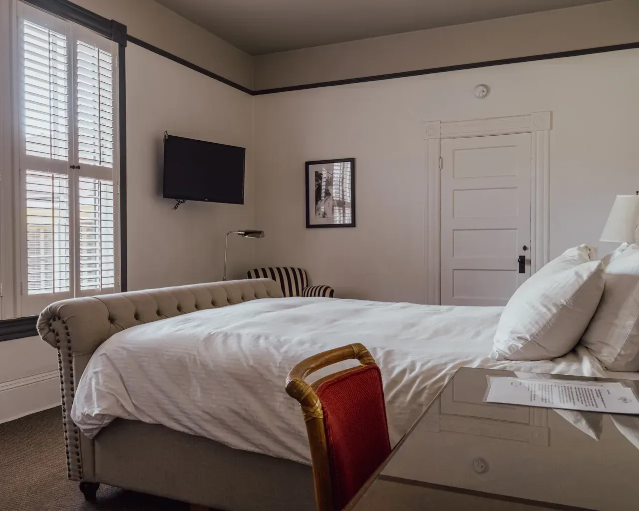 A bedroom with a tufted beige bed, white bedding, a wall-mounted TV, a chair, and a desk against a neutral wall.