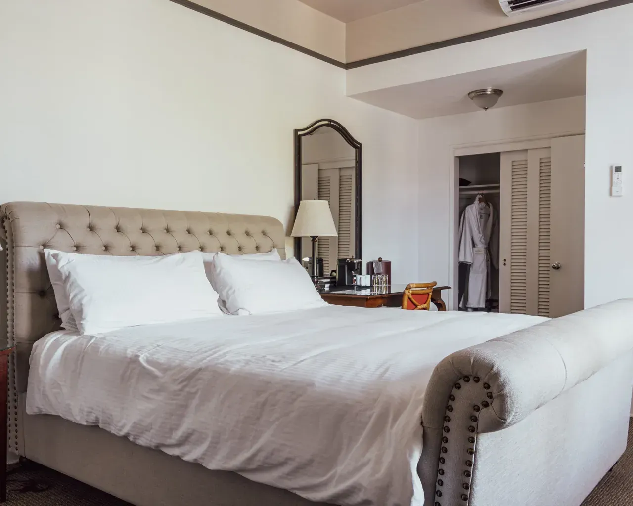 A light-colored, tufted, sleigh-style bed in a hotel room with a desk, mirror, and open closet in the background.