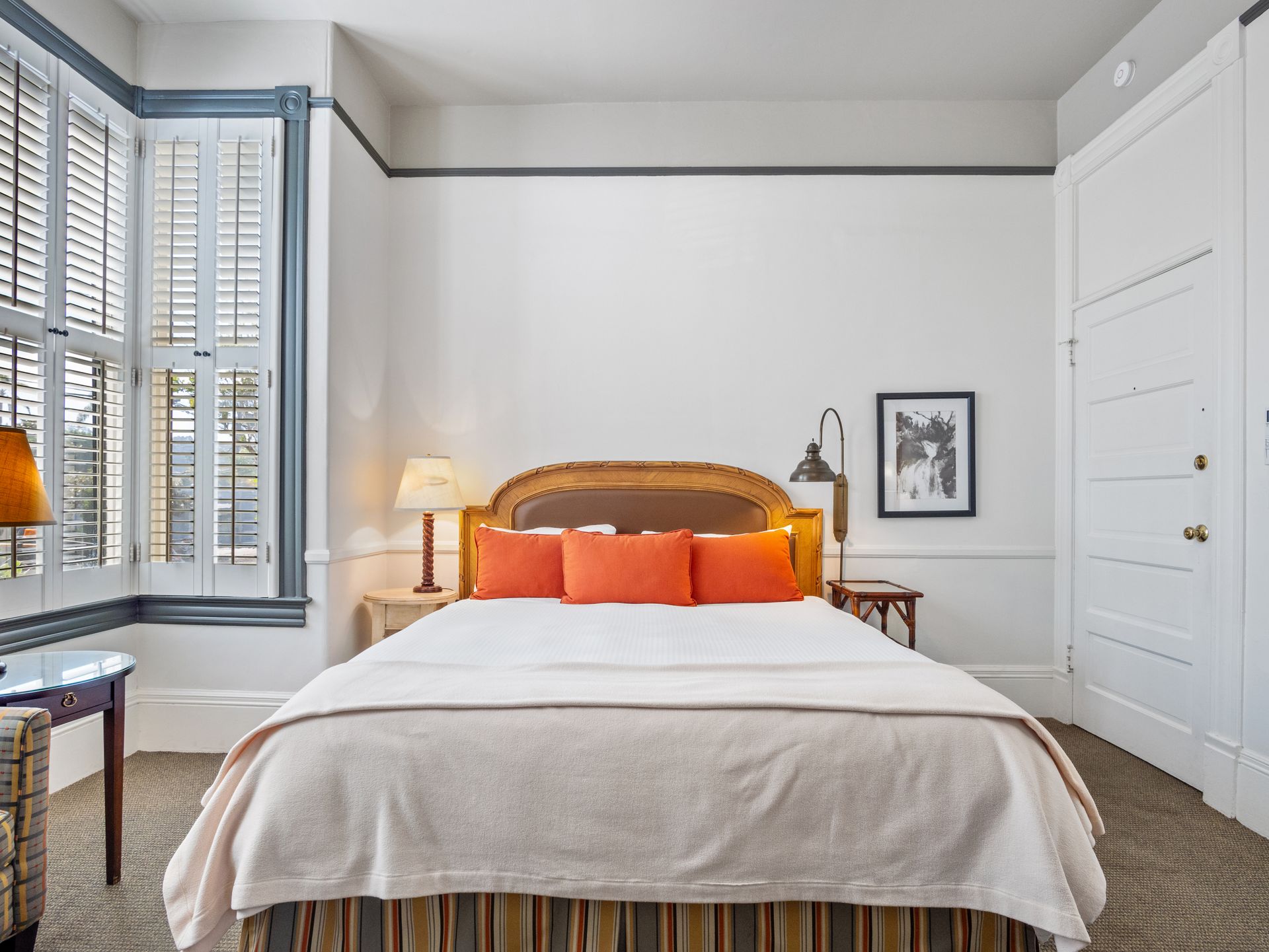 A clean hotel room with a bed centered against a white wall, featuring orange pillows, two lamps, and window shutters.