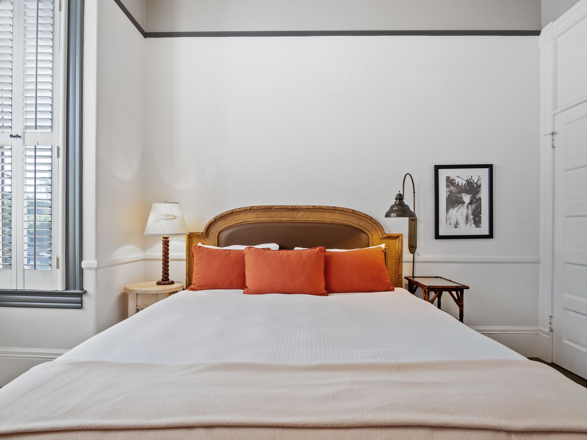 A bed with orange pillows and a gold headboard sits in a white room with a lamp, a side table, and wall art.