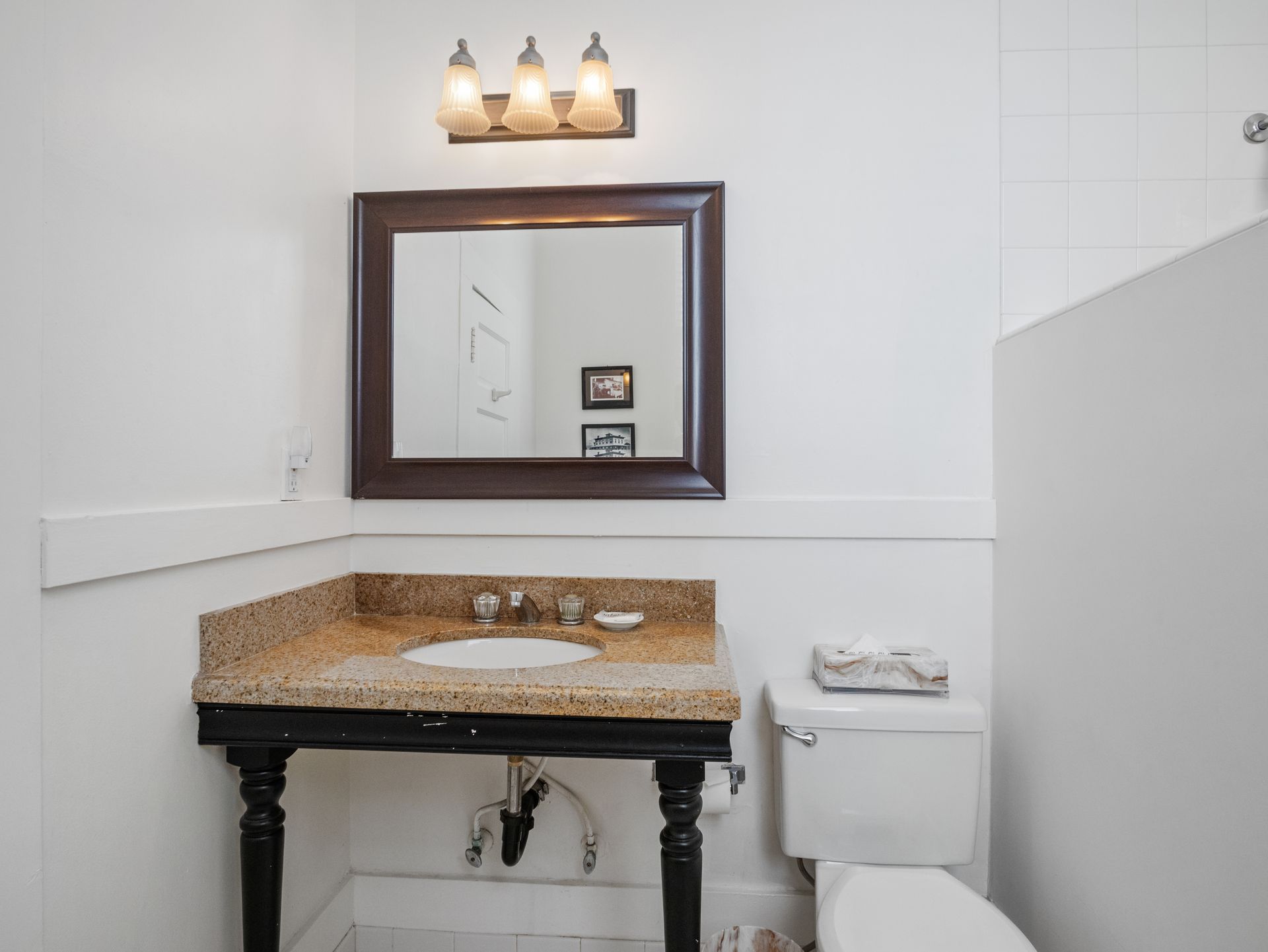 A small bathroom with a granite-topped pedestal vanity, a dark wood-framed mirror, three-bulb vanity light, and toilet.