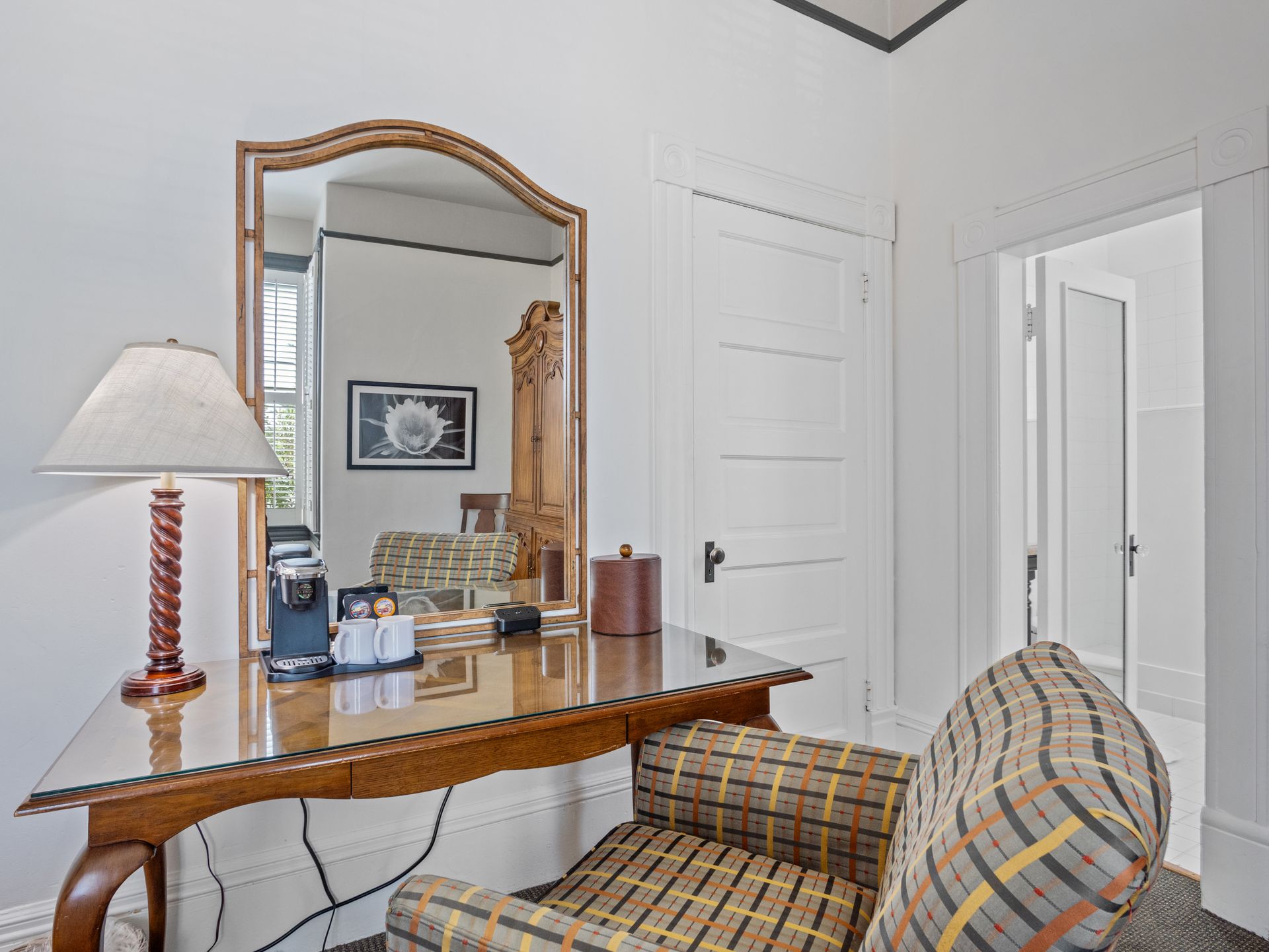 A wooden vanity desk with a glass top, lamp, coffee maker, and mirror, positioned next to a chair in a white room.