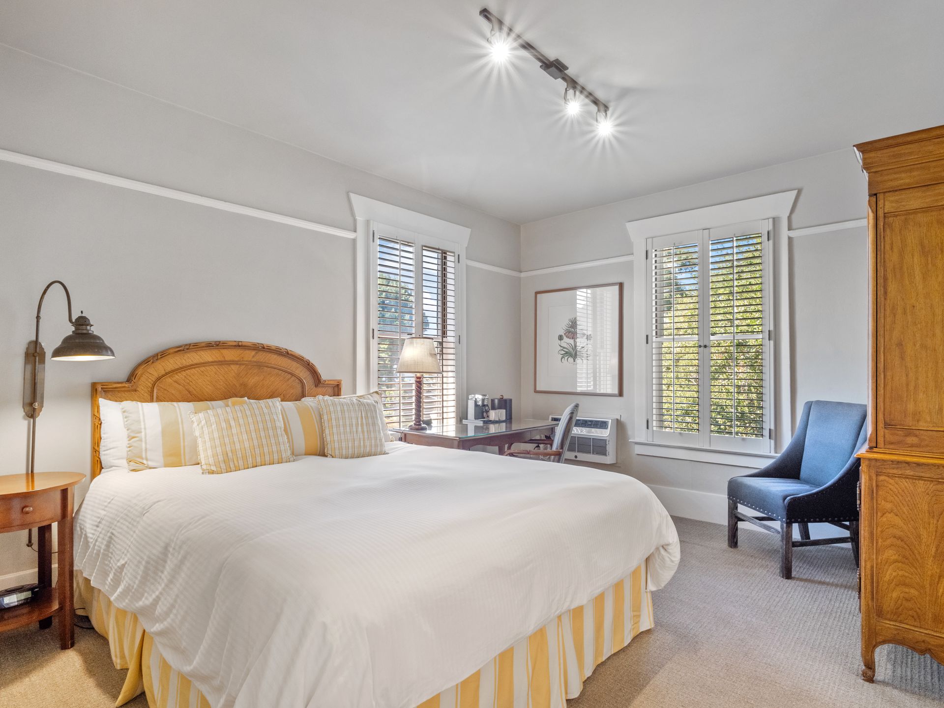 A bedroom with a king-sized bed, a wooden wardrobe, a blue armchair, and large windows with plantation shutters.