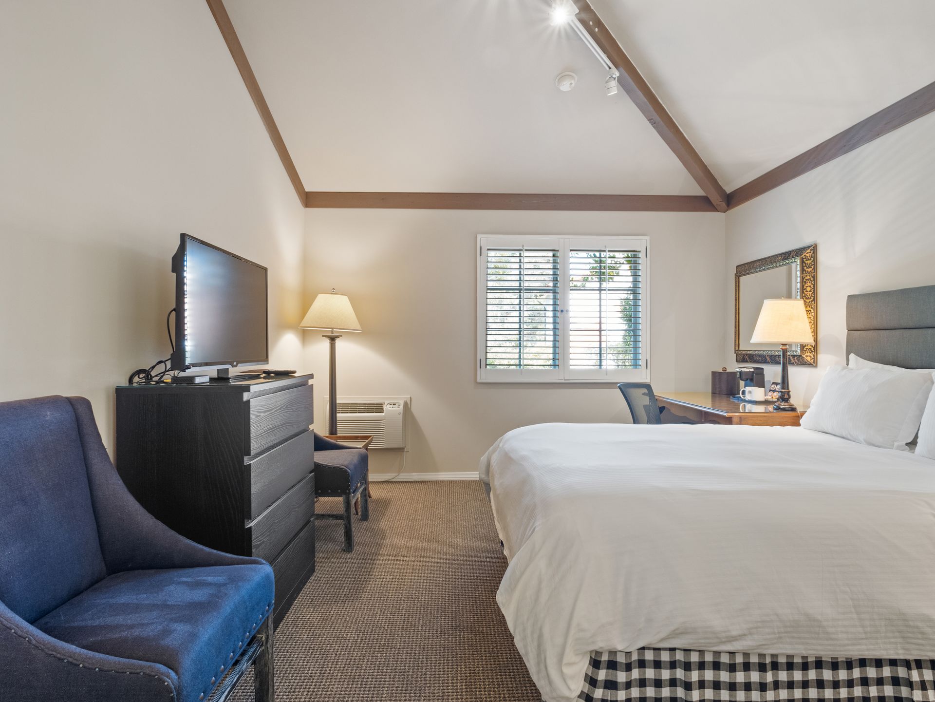 A hotel room with a large bed, a television on a dresser, a blue armchair, a desk, and a window with white shutters.