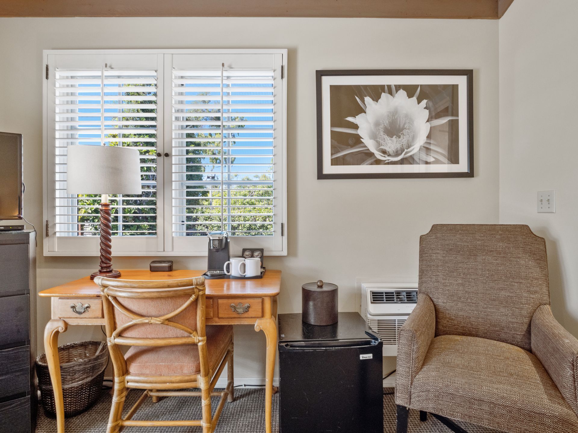 A desk with a lamp, a wicker chair, a small refrigerator, and an armchair next to a window with shutters.