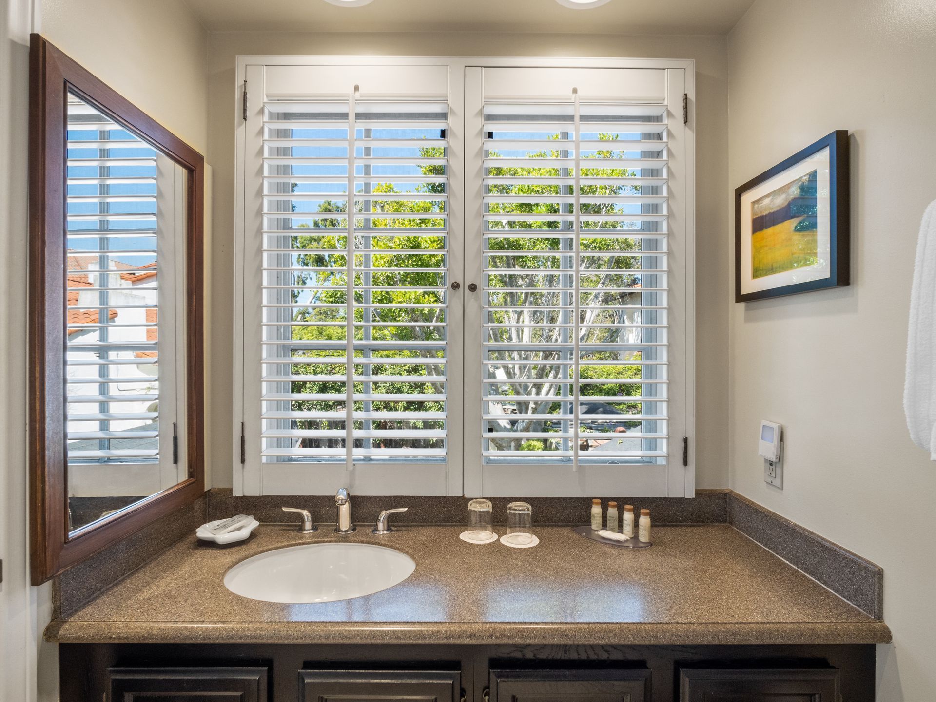 Bathroom vanity with a round sink, stone countertop, plantation shutters over a window, and a wall mirror.