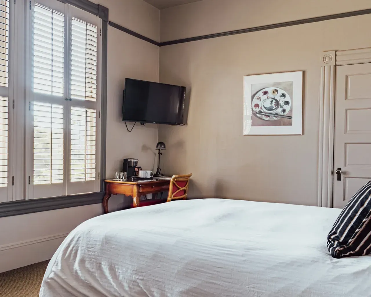 A hotel room featuring a white-bedded bed, a small desk with a coffee maker, a wall-mounted TV, and window shutters.