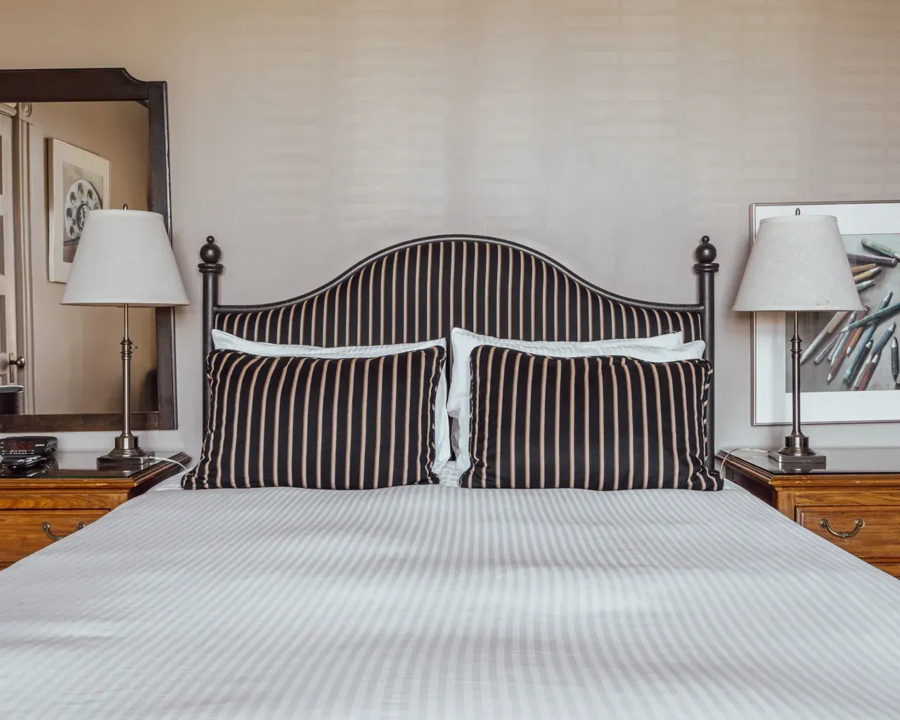 A bed with a dark striped headboard and pillows, flanked by two wooden nightstands with lamps and framed wall art.