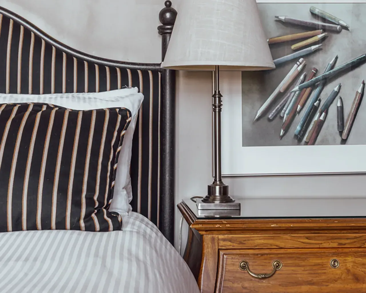 A bed with black-and-tan striped headboard and pillow, next to a wood nightstand with a lamp and framed art of pens.