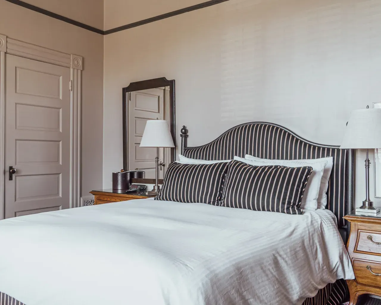 A hotel bedroom featuring a queen bed with a striped headboard, white bedding, and two matching nightstands with lamps.