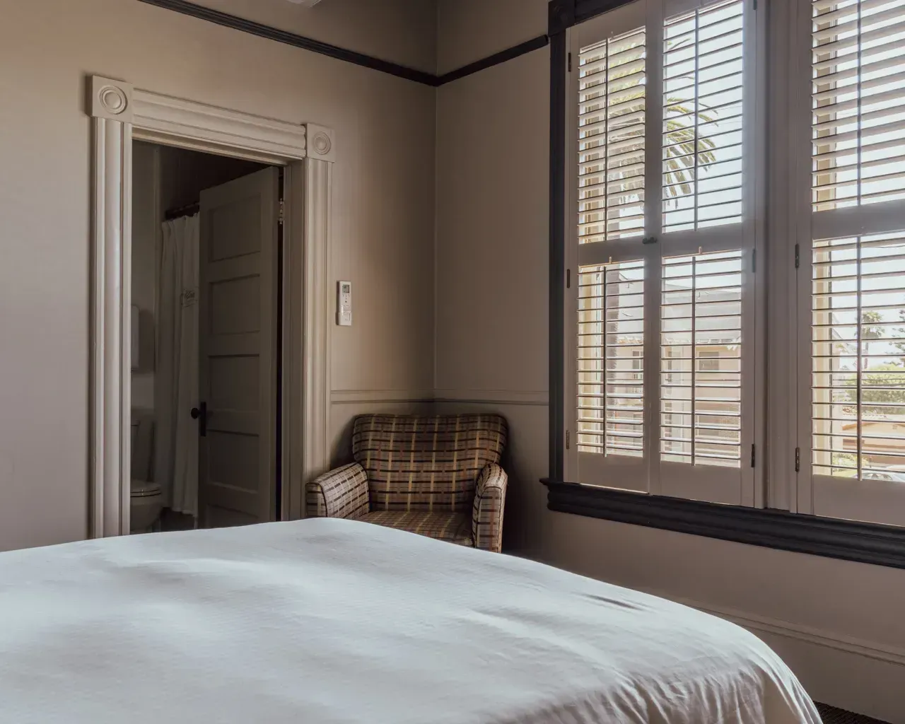 A neutral-toned bedroom with a white bed, a plaid armchair, a doorway leading to a bathroom, and shutters on the window.