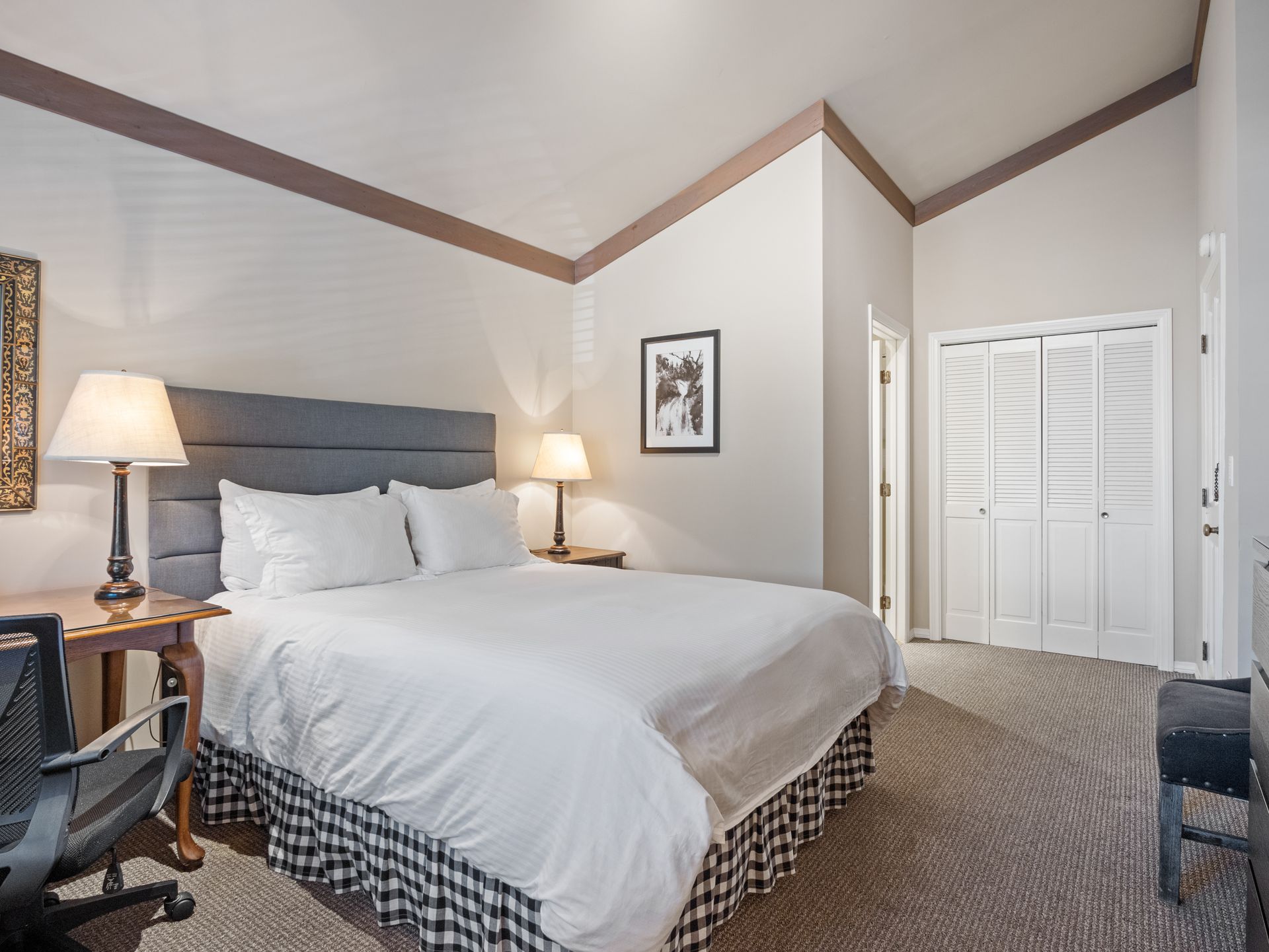 A bedroom with a queen bed featuring a grey headboard, white bedding, and a buffalo check bed skirt, plus a small desk.